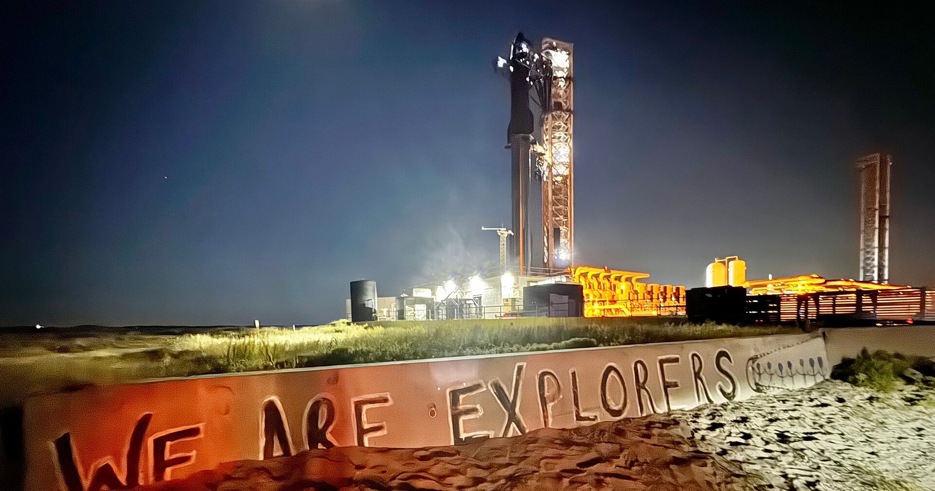 Starship auf der Startrampe in Starbase bei Nacht, davor ein Schriftzug «We are explorers»